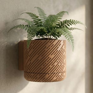 Op de afbeelding: Een lichtbruine, houten wandplanter met een diagonaal streepjespatroon, met een groene varen. De planter is bevestigd aan een bijpassende wandbevestiging. De varen heeft meerdere groene bladeren.