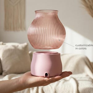 Op de afbeelding: Een tweedelige decoratieve vaas in roze. De bovenkant is een geribbeld, helder glas en de basis is effen, ondoorzichtig roze. De tekst "customizable in colors" is opgenomen. De vaas wordt in de hand van een persoon gehouden.