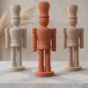 Peut inclure: Trois figurines de casse-noisette debout avec une finition mate. La figurine de gauche est beige clair, celle du centre est orange terre cuite et celle de droite est d'un ton beige sourd. Chaque casse-noisette a un grand chapeau et une base.