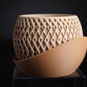 Può includere: Un vaso di ceramica beige con un motivo geometrico a rombi. Il vaso poggia su un sottovaso abbinato. Il design è moderno e il colore è neutro, adatto a vari stili di arredamento. Il vaso è probabilmente per uso interno.