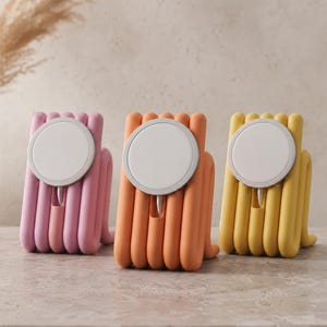Può includere: Tre supporti di ricarica wireless in rosa, arancione e giallo. Ogni supporto presenta un design arrotondato e scanalato con una base di ricarica magnetica bianca circolare montata sul davanti. Un piccolo cavo esce dalla base di ogni caricatore.