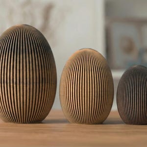 Könnte beinhalten: Drei eiförmige Holzskulpturen mit vertikalen Rillen. Die größte ist hellbraun, die mittlere hat eine ähnliche Farbe und die kleinste ist dunkelbraun. Sie sind auf einer Holzoberfläche angeordnet.