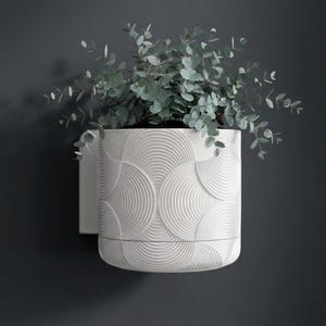 Peut inclure: Un pot de fleurs blanc, mural, avec un motif texturé en forme d'éventail. Le pot cylindrique contient des plantes vertes. Le pot est fixé à un mur gris foncé, créant une esthétique moderne et minimaliste.