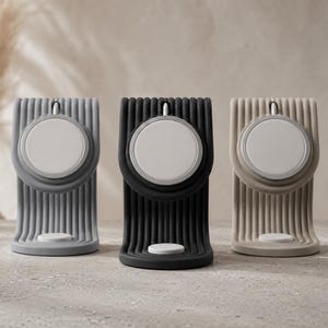 Op de afbeelding: Drie moderne oplaadstandaards in grijs, zwart en beige. Elke standaard heeft een cirkelvormig oplaadpad en een geribbeld, verticaal ontwerp. De standaards zijn ontworpen om een apparaat vast te houden en hebben een minimalistische uitstraling.