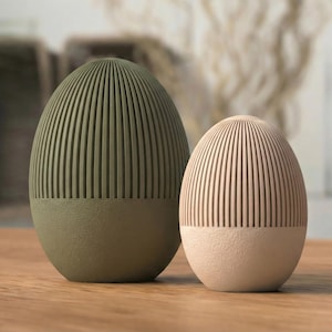 Peut inclure: Deux objets décoratifs en forme d'œuf, l'un vert olive et l'autre rose clair. Les deux ont un motif côtelé sur la moitié supérieure et une couleur unie sur la moitié inférieure. Ils sont posés sur une surface en bois.