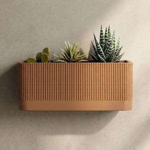 Op de afbeelding: Een rechthoekige, bruine plantenbak met verticale ribbels, met drie verschillende soorten vetplanten. De plantenbak is aan een neutraal gekleurde muur bevestigd, wat een minimalistische esthetiek creëert. De vetplanten omvatten een cactus, een stekelige vetplant en een groen-witte vetplant.
