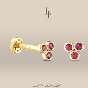 Pode incluir: Um par de brincos de ouro, cada um com três pequenas pedras de rubi dispostas em forma de triângulo.