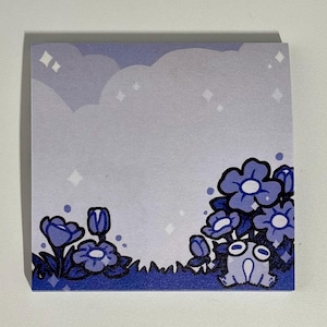 May include: Square sticky note pad with a whimsical design. The pad features a light purple sky with clouds and sparkling stars. The bottom right corner has a cartoon illustration of blue flowers and a small frog.