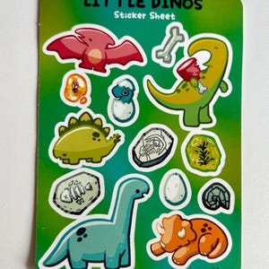 Op de afbeelding: Een stickerblad met een groene achtergrond met verschillende dinosaurus illustraties. Het blad bevat een roze pterodactyl, een groene dinosaurus die een bot eet, een blauwe dinosaurus in een ei, een groene stegosaurus, een blauwe brachiosaurus, een oranje triceratops en verschillende fossiele illustraties.