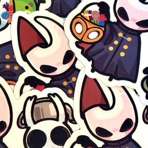 Op de afbeelding: Een verzameling stickers met stripfiguren met bleke gezichten, donkere ogen en bordeauxrode hoorns. De personages dragen donkerblauwe mantels met gouden knopen. Sommigen houden manden vol kleurrijke snoepjes vast.