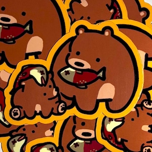 K&ouml;nnte beinhalten: Eine Ansammlung von braunen B&auml;ren-Aufklebern mit gelben Umrandungen. Jeder B&auml;r h&auml;lt einen rot-wei&szlig;en Fisch. Die Aufkleber sind im Cartoon-Stil gehalten.