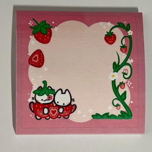 Puede incluir: Un bloc de notas cuadrado rosa con un tema de fresas y gatos. El bloc de notas presenta un borde rosa, un centro rosa claro e ilustraciones de fresas, vides y dos personajes de gatos. Los gatos están dentro de una fresa.