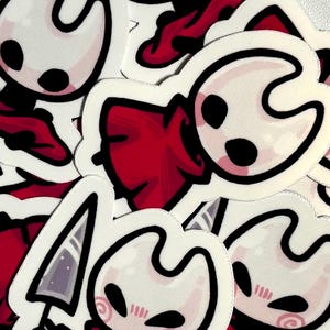 May include: A collection of white stickers featuring a cartoon character with a distinctive head shape, black eyes, and rosy cheeks. The character is wearing a red cloak. Some stickers include a stylized sword. The stickers are on a white background.