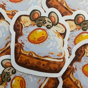 May include: A sticker of a cartoon mouse sleeping on a piece of toast with a fried egg on top. The mouse is brown and has a white belly. The toast is brown and has a yellow egg yolk with white egg whites. The egg yolk has green flecks on it. The sticker is on a brown surface.