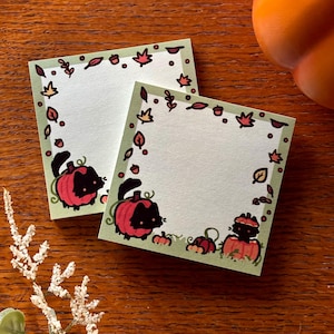 以下が含まれることがあります： 秋の葉とカボチャで飾られた、薄緑色の縁取りのある2つの正方形のメモ帳。各メモ帳には、赤いカボチャの中に黒猫が描かれています。メモ帳は木製の表面にあります。