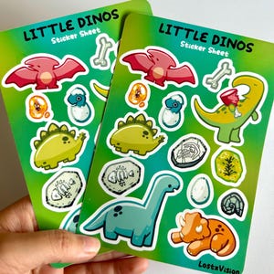 Op de afbeelding: Twee velletjes stickers met een groene achtergrond en kleurrijke dinosaurus illustraties. De velletjes zijn getiteld "Little Dinos Sticker Sheet".