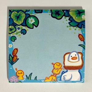 May include: Square notepad with a whimsical illustration of a duck-shaped character in a pond. The border features lily pads and flowers. The duck is depicted with a slice of toast on its head, and there are several smaller ducklings in the water.