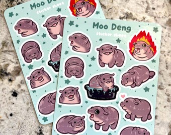 Op de afbeelding: Twee sticker vellen met een lichtblauwe achtergrond en illustraties van cartoon nijlpaarden in verschillende poses. De tekst "Moo Deng sticker sheet" staat op elk vel. De stickers zijn in tinten paars en roze.