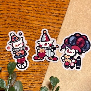 May include: Three die-cut stickers featuring cartoon characters dressed as clowns. One character rides a unicycle, another stands with a ruffled collar, and the third holds a cluster of dark balloons and a balloon animal, all in red, blue, and white.