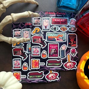May include: A sticker sheet with a black background and a spiderweb pattern. The sheet features various Halloween-themed stickers, including a ghost, a bat, a skeleton fish, a pumpkin pocky, a witch's broom, and a KitKat candy bar. The stickers are in a cartoon style and have a variety of colors, including red, orange, yellow, green, blue, and purple.