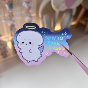 May include: A holographic sticker with a cartoon angel holding a halo. The angel is white with a pink blush and a pink heart on its chest. The sticker has a blue and purple gradient background with stars and hearts. The text on the sticker reads "OMW TO SAVE UR ASS".