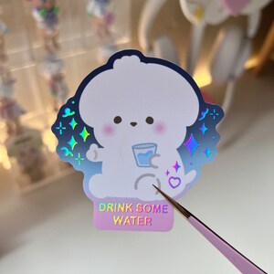 May include: A holographic sticker featuring a white cartoon dog holding a glass of water. The sticker has a blue and purple iridescent background with stars and a heart. The text "Drink Some Water" is printed on the bottom of the sticker.