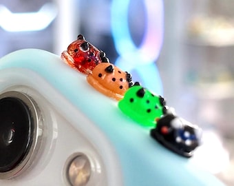 May include: A close-up of a light blue phone case with four colorful frog-shaped charms. The charms are red, orange, green, and black, each with black spots. The phone case has a camera lens.