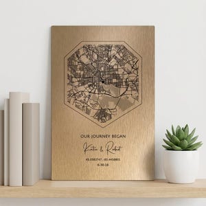 Puede incluir: Una placa rectangular de metal dorado cepillado con un mapa de calles hexagonal negro y un pequeño icono de corazón en el centro. Debajo del mapa, el texto dice: OUR JOURNEY BEGAN, Katie & Robert, 45.0583747, -83.4458801, 6-30-18.