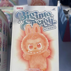 Peut inclure: Une boîte aveugle Pop Mart "Biginto Energy" présente un personnage de lapin de dessin animé avec une fourrure orange et une expression souriante. La boîte a une palette de couleurs bleu clair et orange avec le texte "Biginto Energy" et "The Monsters". La boîte dit aussi "Vinyl Plush Pendant Blind Box".