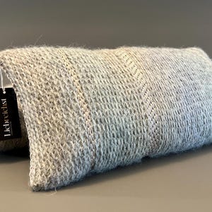 Könnte beinhalten: Zwei rechteckige, hellgraue Strickkissen. Die Kissen haben ein strukturiertes, gewebtes Aussehen und sind übereinander gestapelt. Ein schwarzes Etikett mit dem Text "Liebreichst" hängt am oberen Kissen.