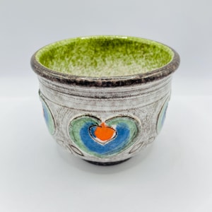 Op de afbeelding: Een keramische plantenbak met een getextureerde, gebroken witte buitenkant en een groene binnenkant. De plantenbak heeft een hartvormig ontwerp in blauw, groen en oranje. De rand is bruin. Deze decoratieve plantenbak is geschikt voor kleine planten.