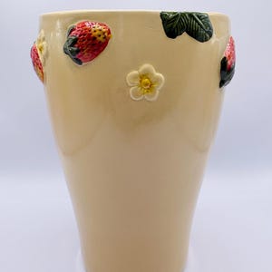 May include: A tall, beige ceramic vase decorated with raised strawberry and flower motifs. The strawberries are red and yellow with green stems, and the flowers are white with yellow centres. The vase has a glossy finish.