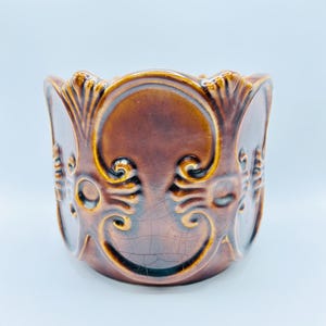 Könnte beinhalten: Ein brauner Keramik-Blumentopf mit glänzendem Finish. Der Topf weist verzierte, erhabene Schnörkel und einen gewellten Rand auf. Das Design umfasst sich wiederholende Muster und eine Craquelé-Textur, die seinen Vintage-Reiz unterstreicht.
