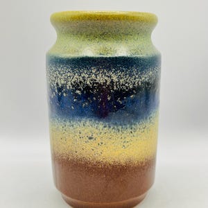 Könnte beinhalten: Eine Keramikvase mit zylindrischer Form und leicht ausgestelltem Rand. Die Vase zeigt einen Farbverlauf, der von Braun am Boden zu Gelb, Blau und Grün an der Oberseite übergeht. Die Oberfläche hat eine gesprenkelte Textur.