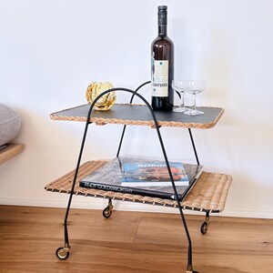 Op de afbeelding: Een vintage serveerwagen met twee niveaus, met een zwart metalen frame en rieten planken. De bovenste plank bevat een wijnfles, twee glazen en een gouden ornament. Tijdschriften liggen op de onderste plank. De wagen heeft kleine gouden wielen.