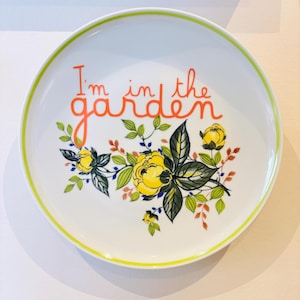 Könnte beinhalten: Weiße Keramikplatte mit gelb-grünem Blumendesign und dem Text "I'm in the garden" in orangefarbener Schrift. Die Platte hat einen dünnen, leuchtend gelben Rand.