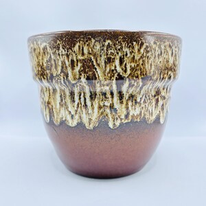 Pode incluir: Um vaso de cerâmica com uma base lisa de cor castanho-escuro sólido que transita para um padrão de esmalte texturizado e escorrido em creme e castanho-escuro ao longo da borda superior. O recipiente tem uma abertura larga e forma arredondada.