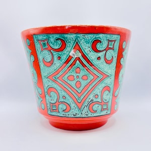Könnte beinhalten: Keramik-Blumentopf mit lebendigem Design. Der Topf hat eine rote Außenseite mit türkisfarbenen geometrischen Mustern und Schnörkeln. Das Design beinhaltet eine zentrale Raute mit einem floralen Motiv. Der Topf hat eine leicht ausgestellte Form und einen abgerundeten Boden.