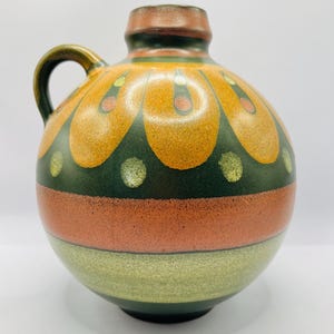 May include: A round, ceramic jug with a handle, featuring a decorative pattern. The jug has a brown top and handle, with a design of orange and green shapes. The body is striped with orange and green bands, creating a vintage aesthetic.