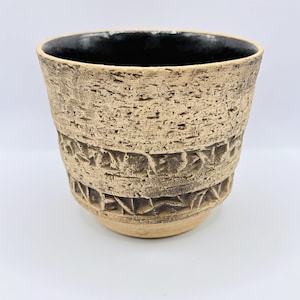 May include: A ceramic cup with a rough, textured beige exterior featuring two horizontal bands of carved geometric patterns. The interior of the cup is finished with a smooth, glossy black glaze, creating a contrast with the earthy, porous-looking outer surface of the vessel.