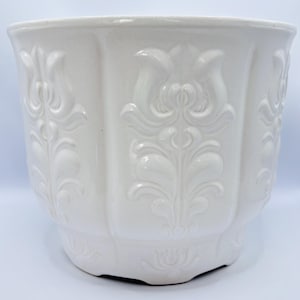 Könnte beinhalten: Weißer Keramik-Blumentopf mit glänzendem Finish. Der Topf hat ein detailliertes geprägtes Blumendesign. Er hat eine breite, abgerundete Form mit leicht ausgestelltem Rand und dekorativem Sockel. Ideal für den Innen- oder Außenbereich.