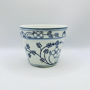 Op de afbeelding: Een witte keramische bloempot met een handgeschilderd blauw bloemmotief. Het ontwerp bevat delicate ranken, bladeren en cirkelvormige bloesems rondom de buitenkant, met horizontale blauwe banden langs de rand en de onderkant van de pot.