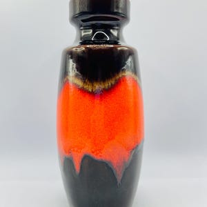 Puede incluir: Un jarrón de cerámica alto con acabado brillante. El jarrón presenta una llamativa combinación de colores, con una sección central naranja vibrante, que pasa a una parte superior e inferior marrón oscuro. El diseño tiene una estética moderna de mediados de siglo.