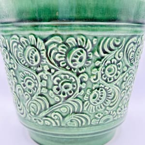 Könnte beinhalten: Ein grüner Keramik-Blumentopf mit detailliertem Blumenmuster. Der Topf hat einen breiten Rand und einen leicht konischen Körper. Das geprägte Design zeigt stilisierte Blumen und Blätter, die Textur hinzufügen. Geeignet für den Innen- oder Außenbereich.
