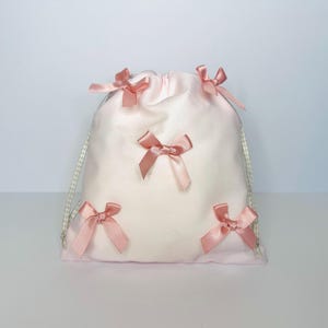 Puede incluir: Una bolsa de cordón rosa con cinco lazos de satén rosa. La bolsa tiene un patrón de cuadros vichy blanco y gris en el cordón.