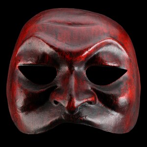 May include: A full-face mask featuring a deep red and black mottled finish. The mask displays a prominent brow ridge, a defined nose, and two wide, almond-shaped eye openings. The surface texture appears smooth with subtle variations in colour.