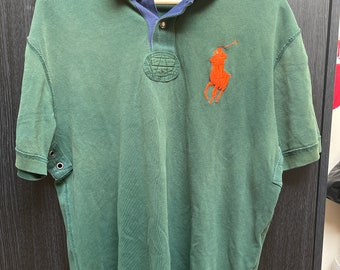 Peut inclure: Polo vert avec un col bleu et jaune, un logo de joueur de polo brodé en rouge et un patch circulaire sur la poitrine. Le polo a un col boutonné et des manches courtes.