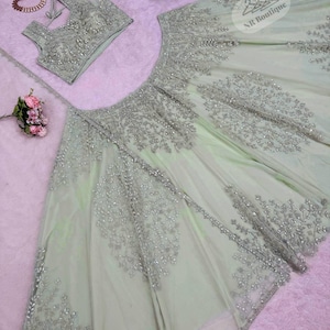 May include: A light green, intricately embroidered formal dress set. The set includes a sleeveless top, a full skirt, and a matching sheer scarf. The embroidery is silver and features floral patterns. The set is displayed on a pink surface.