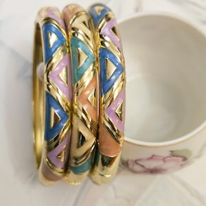 May include: Three gold bangles with colorful enamel geometric designs in blue, pink, green, and peach. The bracelets are stacked together, showcasing their vibrant colors and intricate patterns. A white teacup with a floral design is in the background.