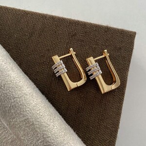 Könnte beinhalten: Ein Paar goldfarbene Creolen mit rechteckigem Design. Jeder Ohrring hat einen kleinen Abschnitt mit drei Reihen weißer Steine. Die Ohrringe werden auf einer braunen Oberfläche präsentiert.